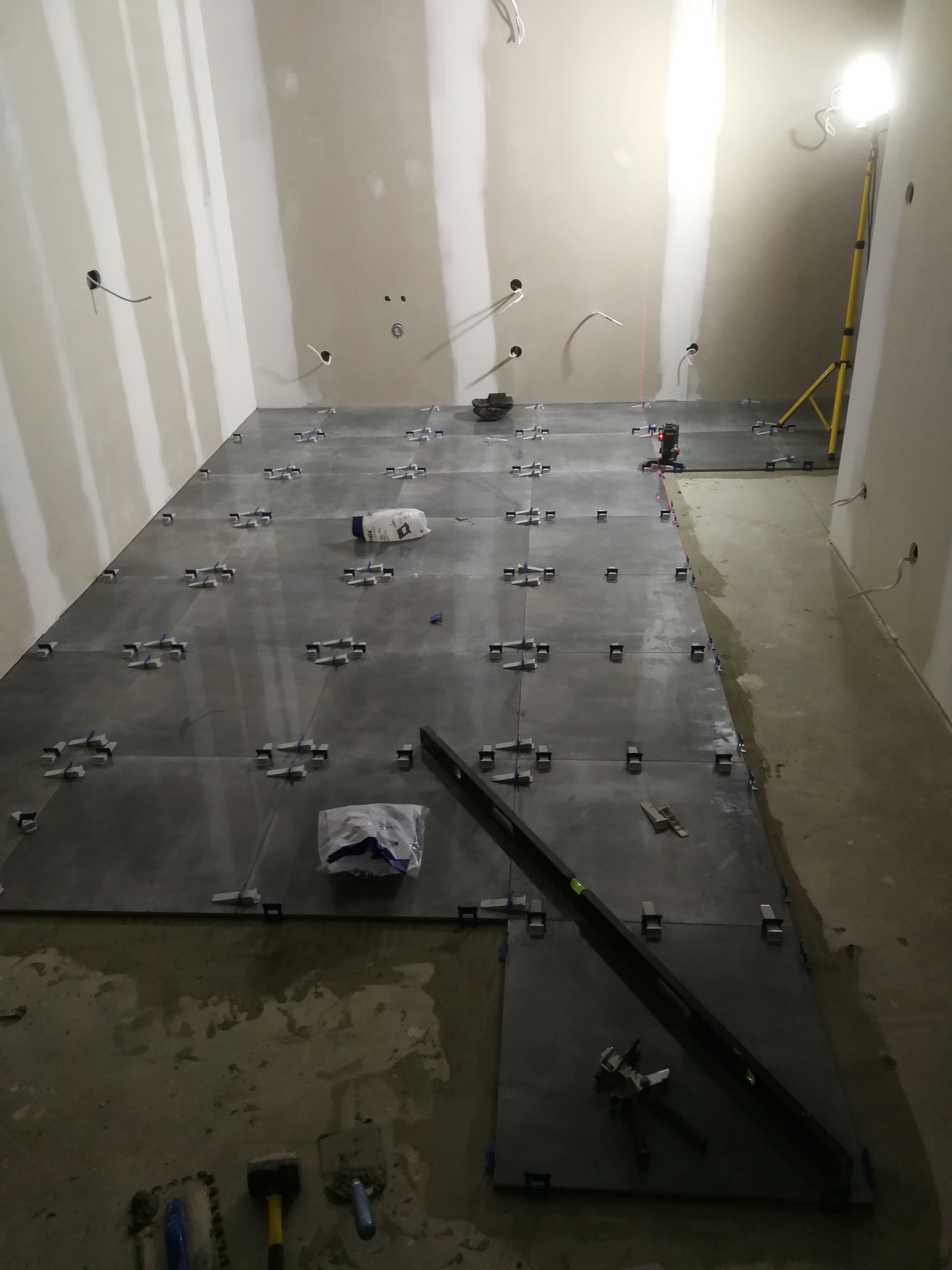Układanie ciemnoszarych płytek na podłodze w surowym pomieszczeniu, widoczne poziomica, paczki z klejem i narzędzia, laser krzyżowy wyznacza linię, oświetlenie budowlane