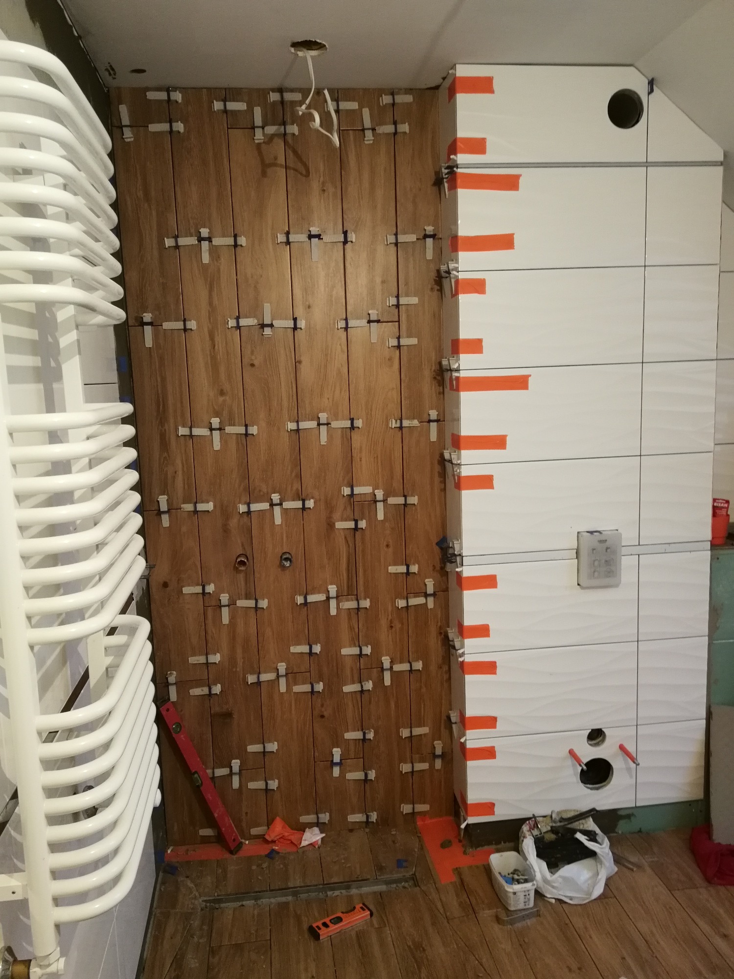 Układanie płytek imitujących drewno na ścianie łazienki, widoczne krzyżyki dystansowe i poziomica, fragment grzejnika drabinkowego.