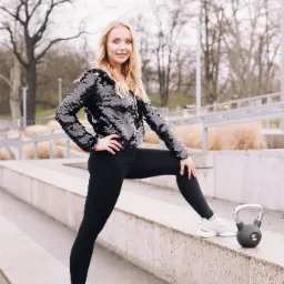 Blondynka w cekinowej bluzie, czarnych legginsach i białych butach sportowych, opiera nogę na betonowym murku obok czarnego kettlebella 16 kg, w tle park.