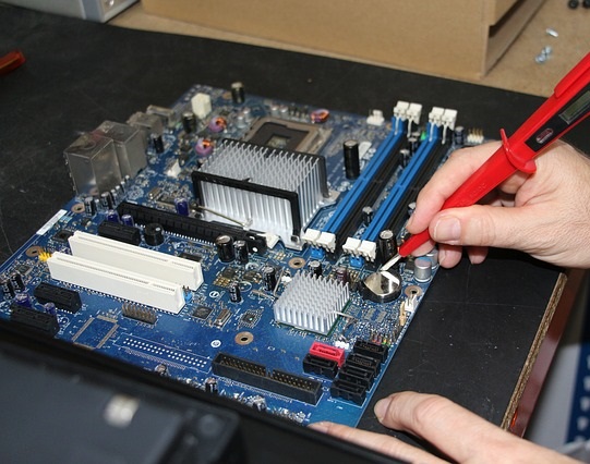 Diagnostyka płyty głównej komputera: ręka z czerwonym multimetrem mierzy napięcie baterii CMOS na niebieskiej płycie głównej z radiatorami i slotami pamięci RAM.