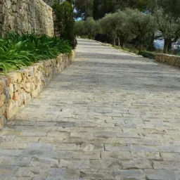 Długa, wyłożona jasną kostką droga wznosi się łagodnie pod górę, ograniczona z obu stron niskimi murkami z kamienia, na których rosną zielone rośliny. W tle widoczne drzewa i zabudowania.