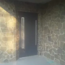 Wejście do budynku obłożone nieregularnymi płytami kamiennymi w odcieniach szarości i beżu, z nowoczesnymi, antracytowymi drzwiami z pionowym przeszkleniem.