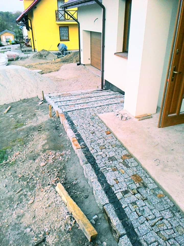 Ścieżka z kostki granitowej w trakcie budowy przy domu, widoczne przygotowane podłoże, krawężniki i częściowo ułożona nawierzchnia z kostki, z elementami w kolorze rdzawym.