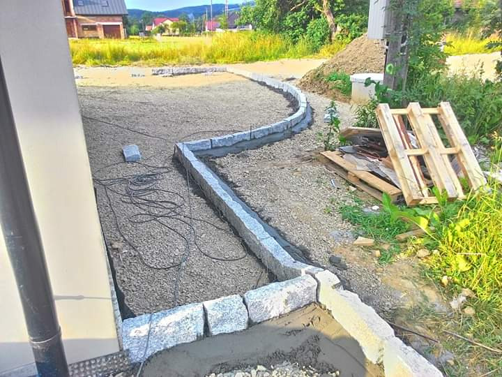 Krawężnik z szarej kostki granitowej układany na podsypce cementowej, widoczne palety i przygotowany teren pod przyszły chodnik.