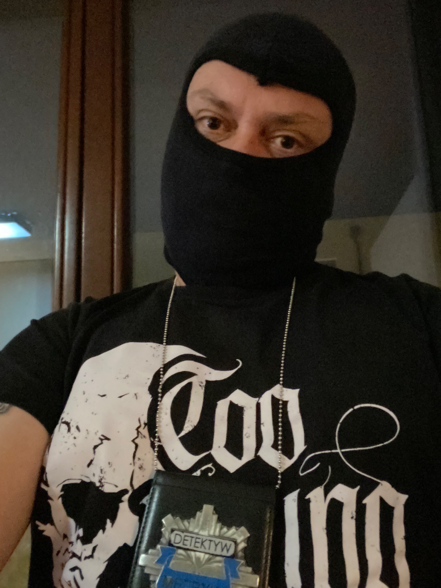 Osoba w czarnej kominiarce i koszulce z nadrukiem, nosząca identyfikator z napisem 'Detektyw' na łańcuszku.