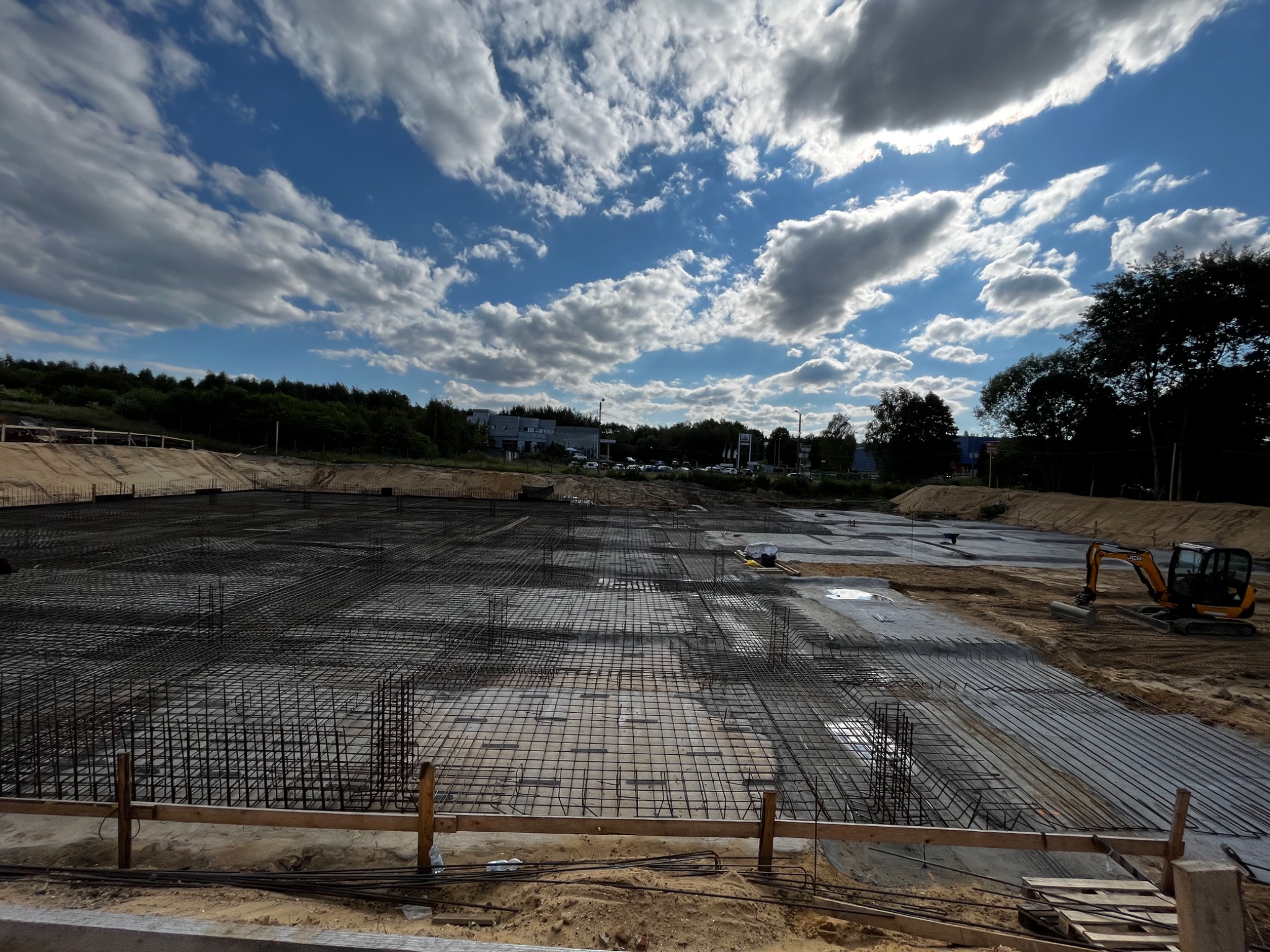 Szeroki kadr na przygotowany teren pod budowę z siatką zbrojeniową, częściowo zalany betonem, z małą koparką JCB w prawym rogu, pod błękitnym niebem z białymi chmurami.