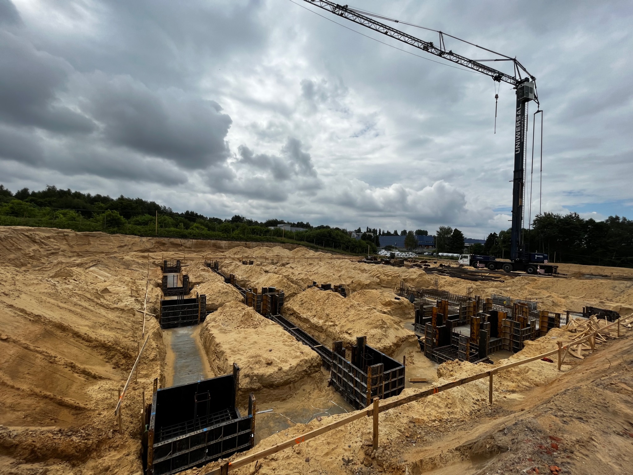 Wykop pod fundamenty z szalunkami i widocznym zbrojeniem, na placu budowy stoi żuraw, w tle zalesiony teren i pochmurne niebo.