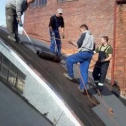 Czterech dekarzy na skośnym dachu podczas remontu z użyciem papy termozgrzewalnej, widoczny budynek z czerwonej cegły w tle.
