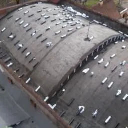 Dach pokryty papą termozgrzewalną z widocznymi zakładkami i fragmentami niezabezpieczonymi, ujęcie z góry, widoczne otoczenie miejskie.