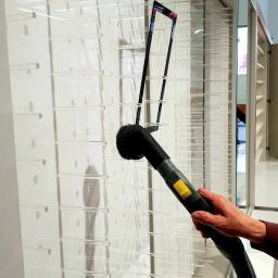 Sprzątanie24 Natalia Lamentowicz - Osoba czyści szklaną ściankę z półkami za pomocą odkurzacza z okrągłą szczotką, widoczne elementy konstrukcyjne, oświetlenie wnętrza.