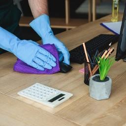 Sprzątanie24 Natalia Lamentowicz - Osoba w niebieskich rękawicach czyści biurko z kalkulatorem, monitorem, klawiaturą i przyborami do pisania, używając fioletowej ściereczki z mikrofibry.