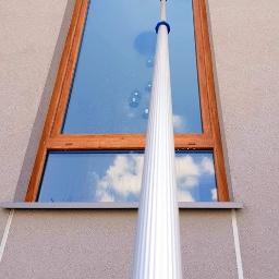 Sprzątanie24 Natalia Lamentowicz - Zewnętrzne okno z drewnianą ramą czyszczone teleskopowym mopem, na szybie widoczne krople i odbicie nieba.