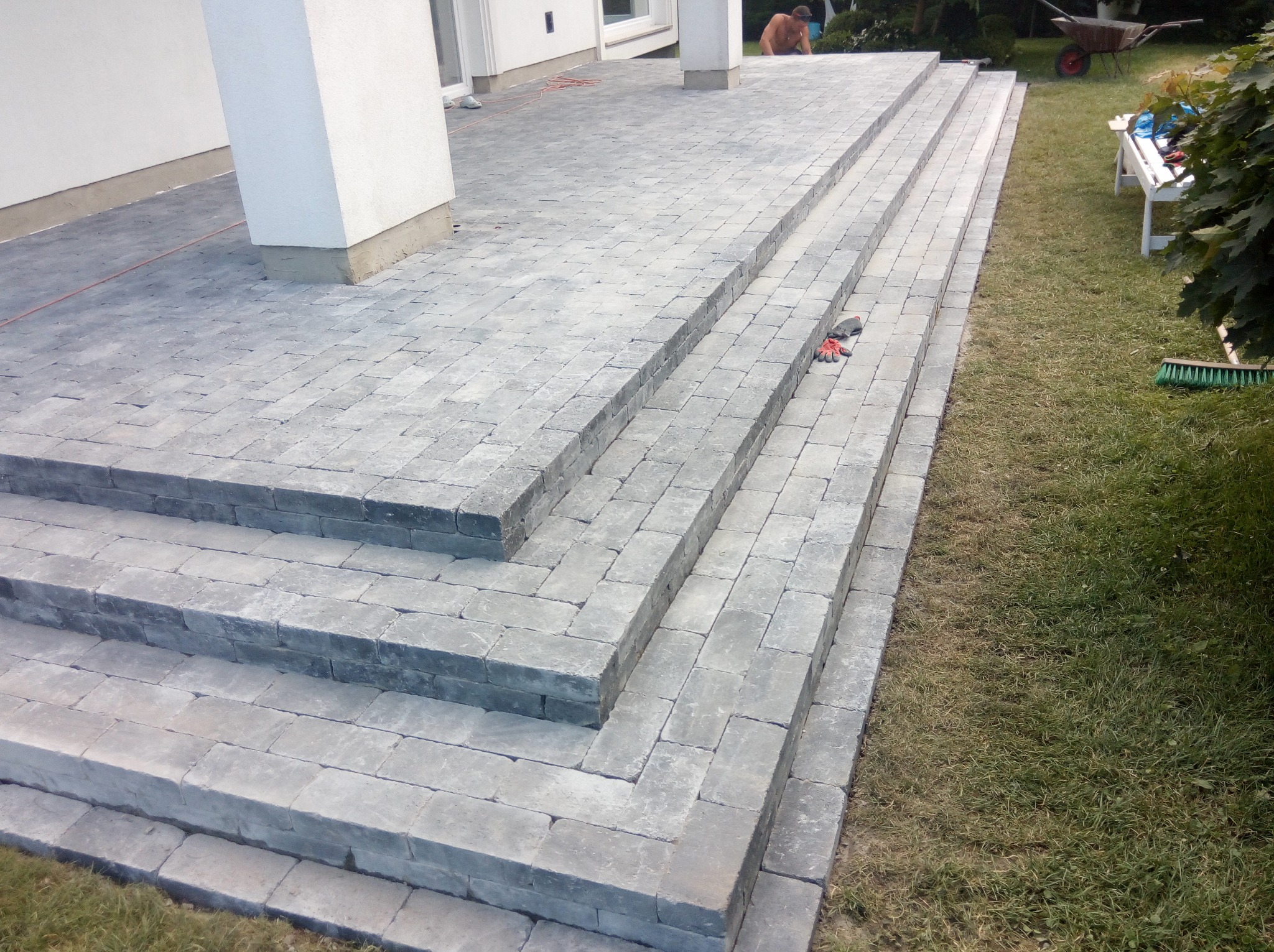 Ułożona kostka brukowa tworząca schody prowadzące do domu, z widocznymi rękawicami roboczymi i fragmentem trawnika