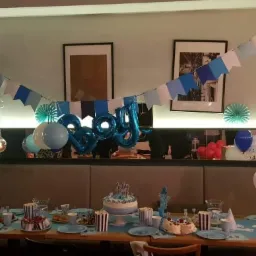 Udekorowany stół na przyjęcie urodzinowe dla chłopca, z tortem, balonami w odcieniach niebieskiego i białego, girlandami z chorągiewek i papierowymi dekoracjami.