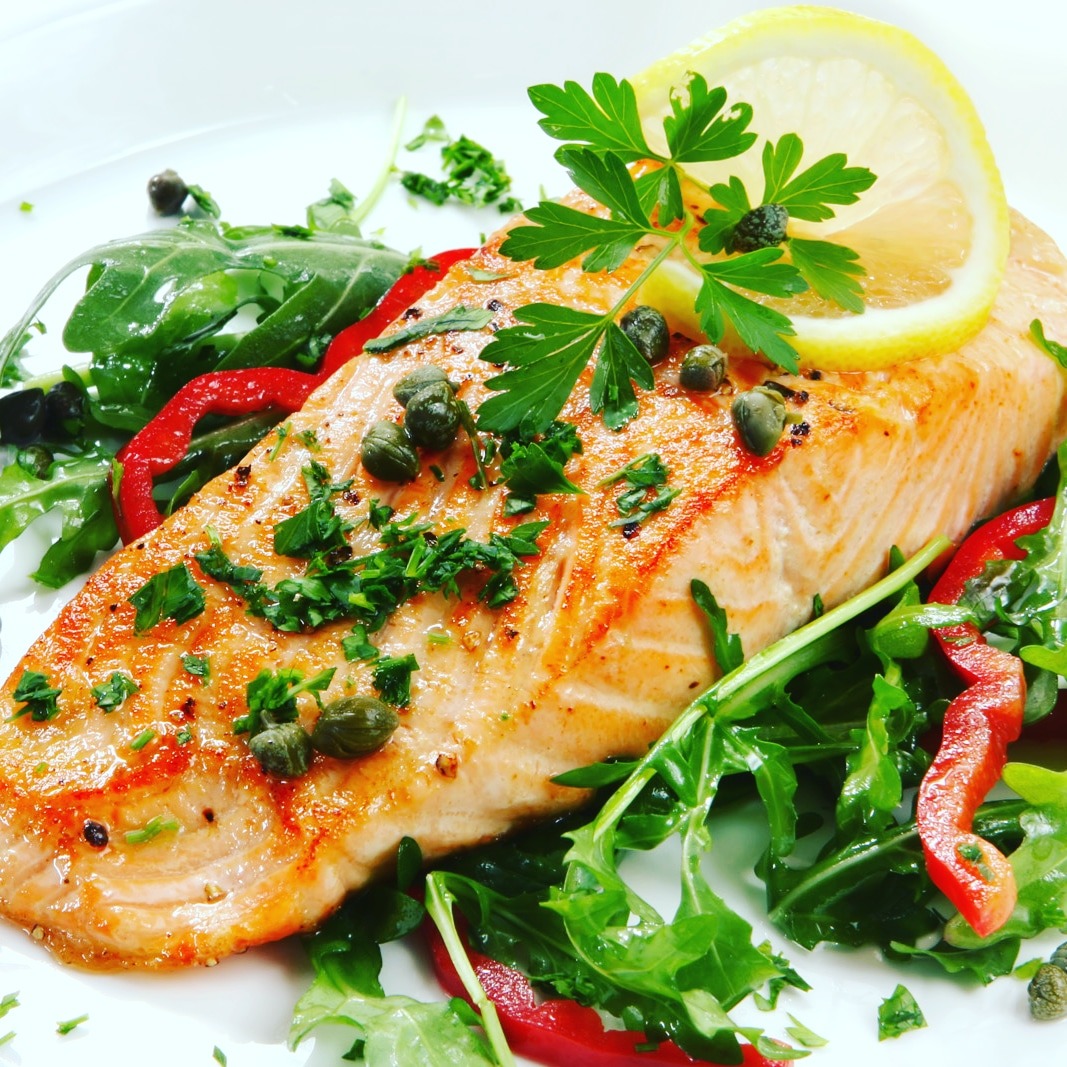 Grillowany filet z łososia na rukoli z czerwoną papryką, kaparami, natką pietruszki i plasterkiem cytryny na białym talerzu.