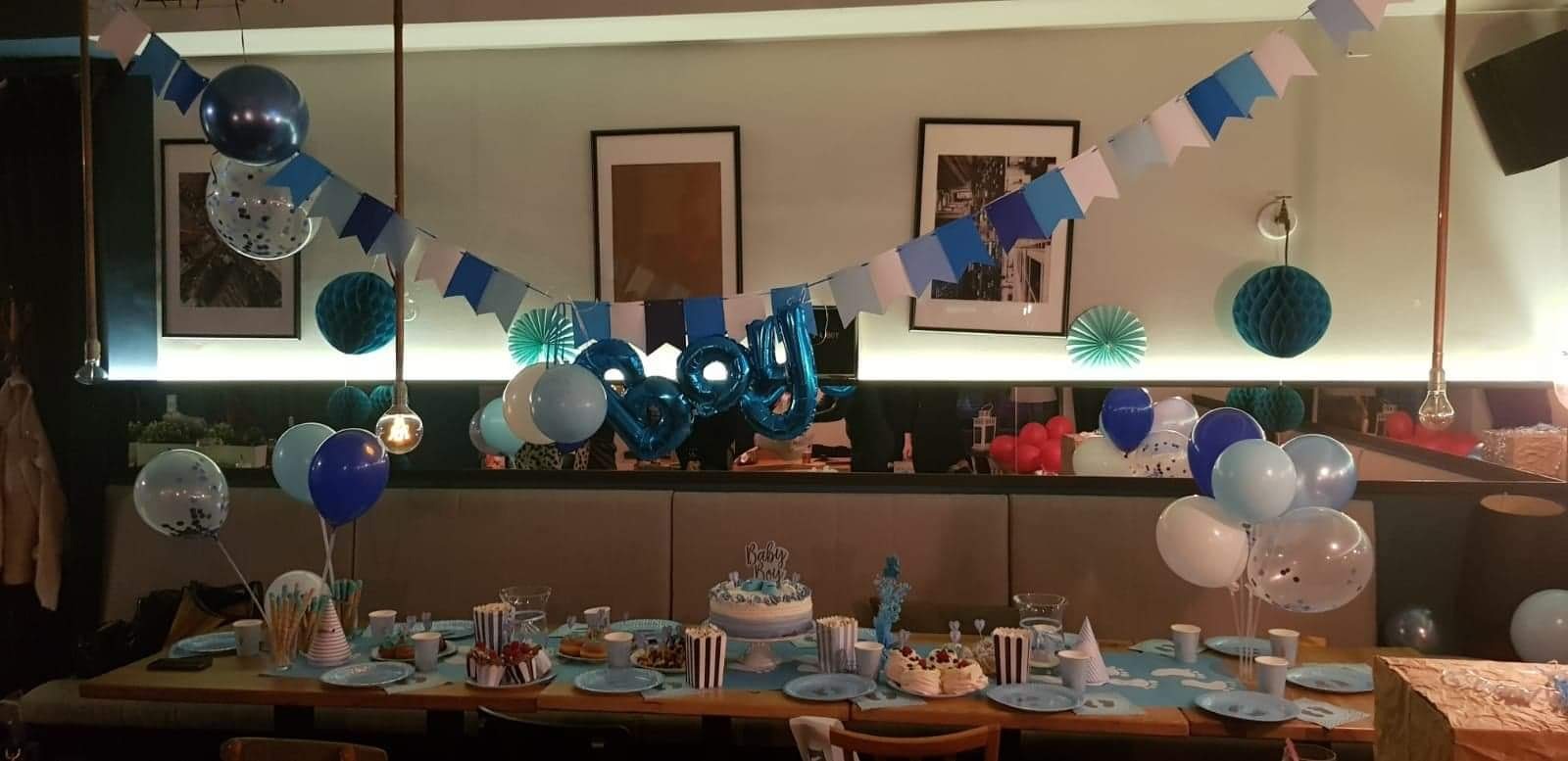 Udekorowany stół na przyjęcie urodzinowe dla chłopca, z tortem, balonami w odcieniach niebieskiego i białego, girlandami z chorągiewek i papierowymi dekoracjami.