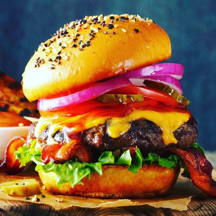 Apetyczny hamburger z wołowiną, bekonem, serem cheddar, pomidorem, ogórkiem kiszonym, czerwoną cebulą i sałatą, podany na drewnianej desce.