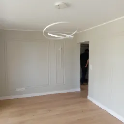 Jasne wnętrze po remoncie: panele podłogowe, ściany z dekoracyjnymi ramami, nowoczesna lampa. W tle osoba przechodząca przez drzwi.