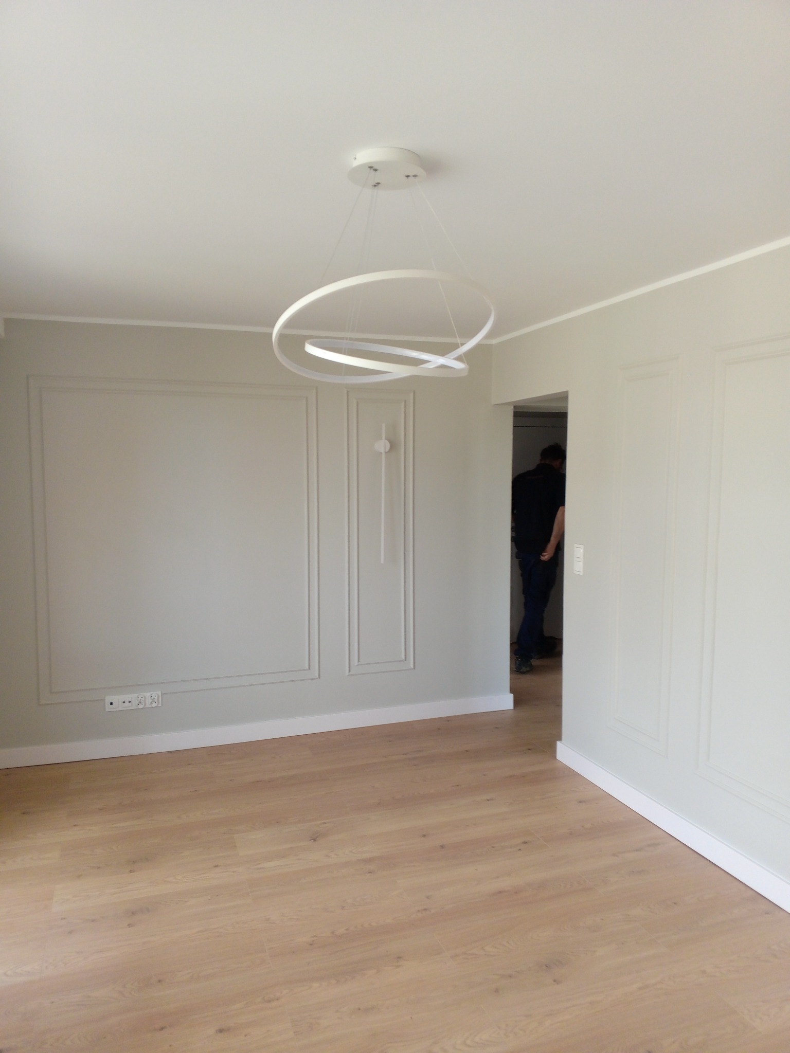 Jasne wnętrze po remoncie: panele podłogowe, ściany z dekoracyjnymi ramami, nowoczesna lampa. W tle osoba przechodząca przez drzwi.