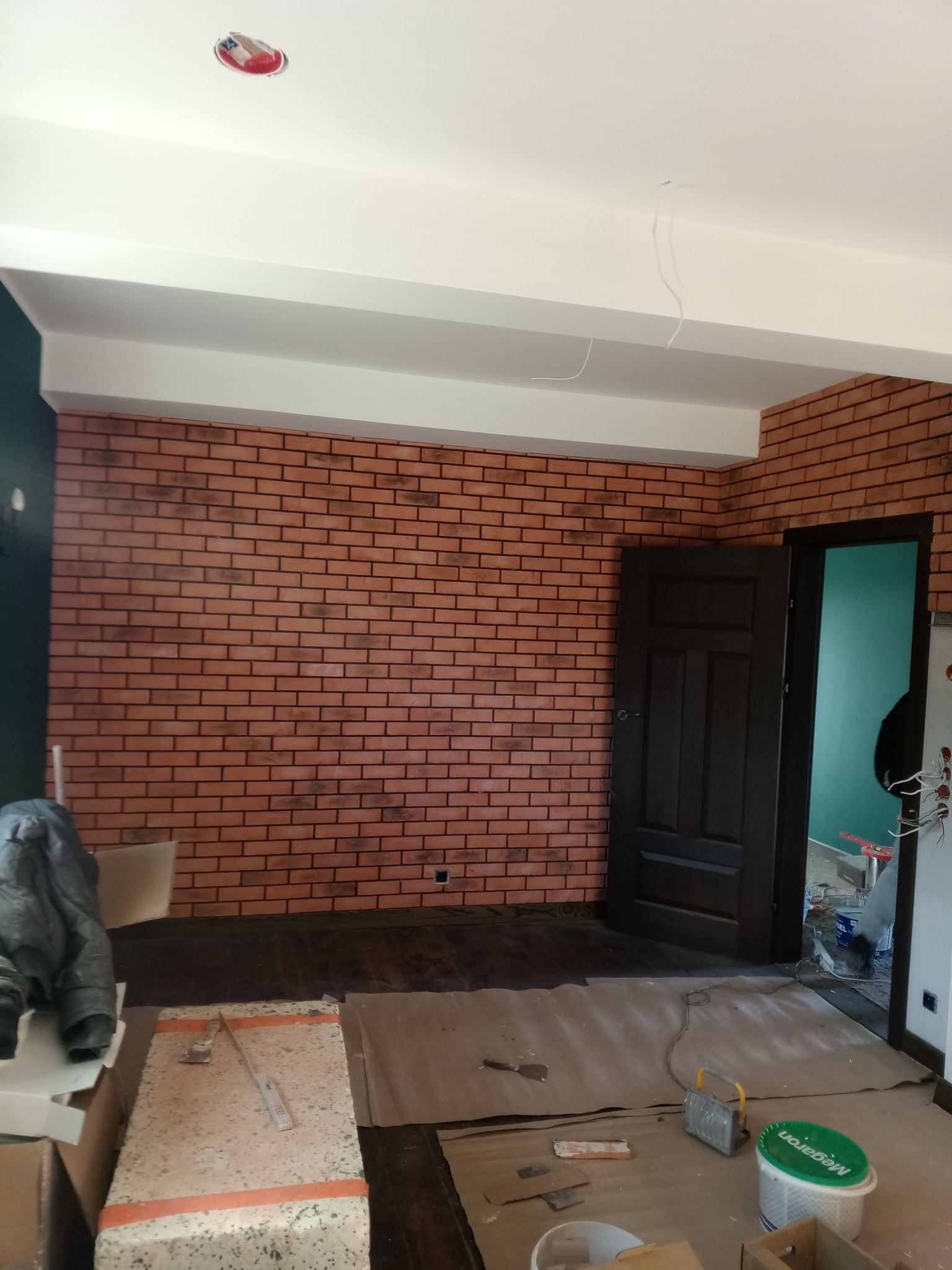Wnętrze z dekoracyjną ścianą z cegły, otwarte drzwi i widoczne narzędzia pracy, sugerujące etap wykończenia. Oświetlenie sufitowe w trakcie montażu.