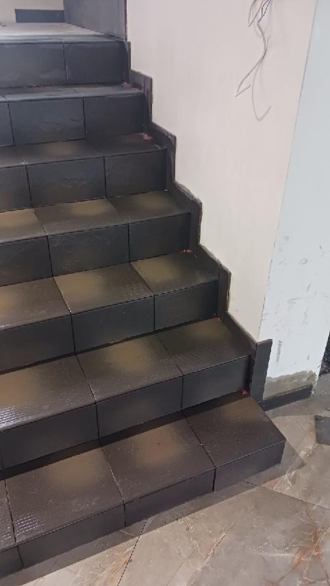 Wykończone schody z ciemnych, kwadratowych płytek, z widocznymi krzyżykami dystansowymi. Ściana obok schodów w trakcie wykończenia.