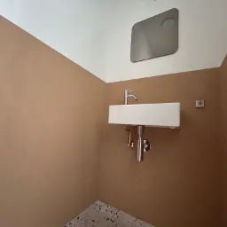 Nowoczesna, prostokątna umywalka z chromowaną baterią i syfonem, zamontowana na ścianie pomalowanej w dwóch odcieniach brązu i bieli, z minimalistycznym lustrem w tle i fragmentem podłogi z lastryko.