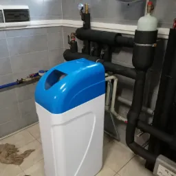Kompaktowy zmiękczacz wody z niebieską pokrywą, zainstalowany w pomieszczeniu z szarymi płytkami, widoczne rury instalacji wodnej i narzędzia na podłodze.