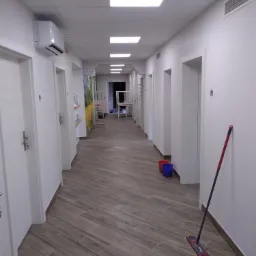 Długi korytarz biurowy z białymi ścianami i drewnopodobną podłogą z płytek, widoczne drzwi, klimatyzacja, mop i dwa wiadra w tle, po remoncie, czysty i jasny.