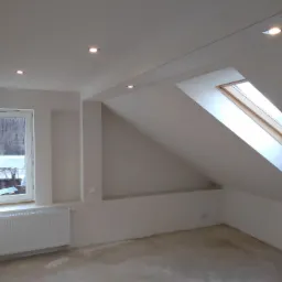 Poddasze po pracach przygotowawczych: ściany i sufit pokryte jednolitą, jasną warstwą, widać okno dachowe i okno w ścianie szczytowej, grzejnik, surowa podłoga przed dalszym wykończeniem.