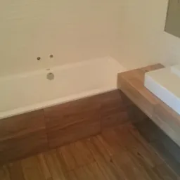 Nowoczesna łazienka w trakcie wykańczania: biała wanna obudowana panelami imitującymi drewno, umywalka nablatowa na drewnianym blacie, białe płytki ścienne z delikatną fakturą, widoczne ślady prac...