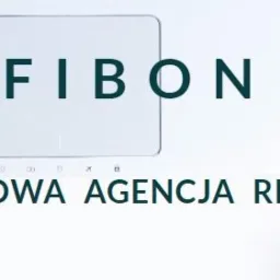 Minimalistyczne ujęcie biurka z widocznym logo 'FIBON Kompleksowa Agencja Reklamowa' nad białym touchpadem i ergonomiczną myszką.