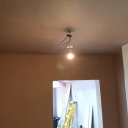 Pokój w trakcie remontu: ściany i sufit pokryte świeżą, brązową warstwą, widoczna goła żarówka na suficie i drabina w przejściu do innego pomieszczenia.