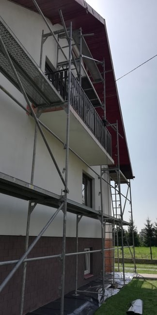 Elewacja domu jednorodzinnego w trakcie prac wykończeniowych, widoczne rusztowania, fragment balkonu z czarną balustradą oraz dolna część elewacji wykończona brązową okładziną.