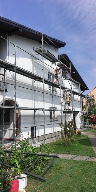 Dwóch mężczyzn pracuje na rusztowaniu przy białym domu, jeden z nich w krótkich spodenkach, drugi w żółtej koszulce, wokół domu zielony trawnik i roślinność, w tle widoczny fragment sąsiedniego...