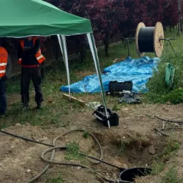 Prace ziemne związane z instalacją kabla telekomunikacyjnego; widoczni pracownicy w kamizelkach odblaskowych pod zielonym namiotem, bęben z kablem, wykop, narzędzia i niebieska plandeka.