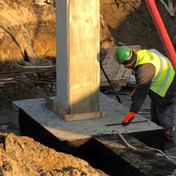 Rathcom - Pracownik w kasku i kamizelce odblaskowej przy betonowym słupie na fundamencie, przygotowujący zbrojenie, widok na wykop i teren budowy w słoneczny dzień.