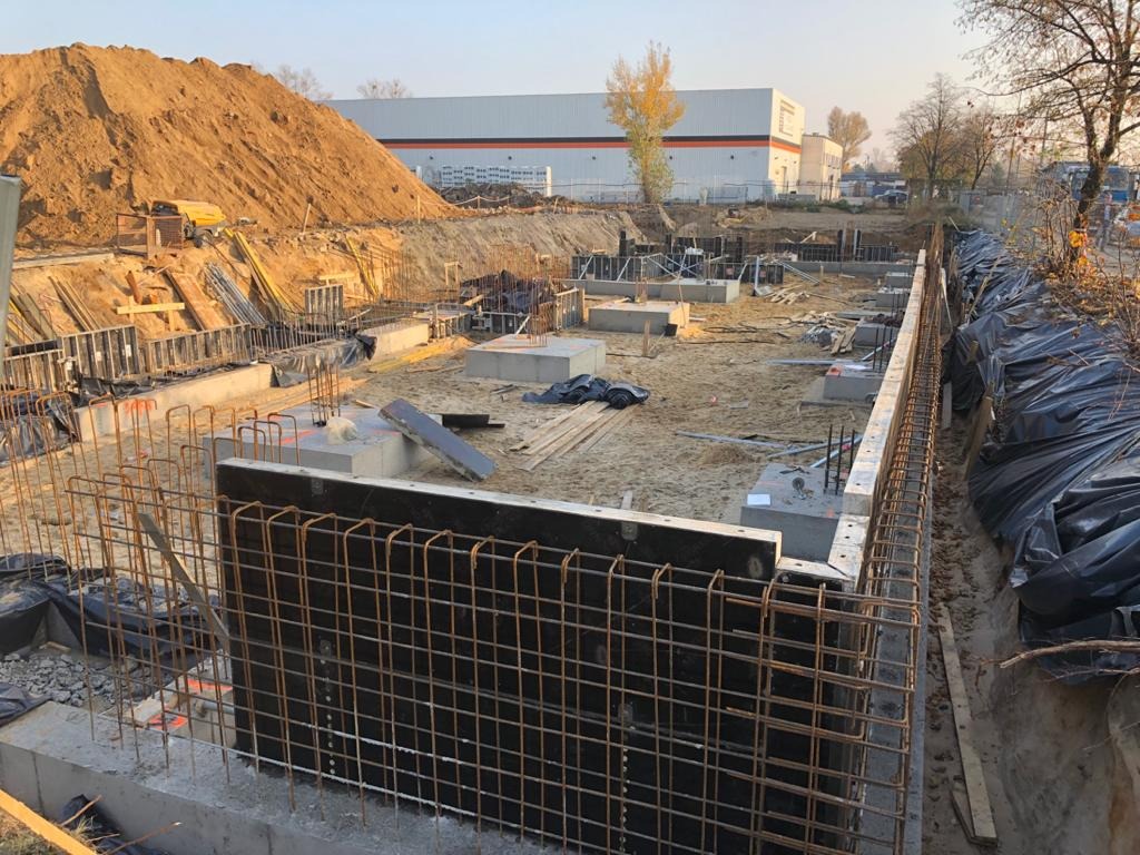 Fundamenty budynku w trakcie budowy, widoczne zbrojenia, betonowe bloczki i duża hałda ziemi w tle.