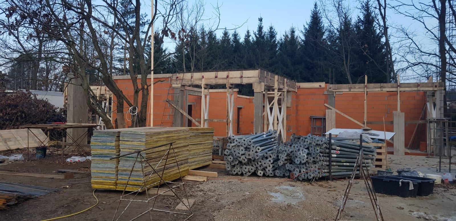 Wznoszenie murów domu z czerwonej cegły, widoczne szalunki drewniane, stemple budowlane i stos płyt szalunkowych na placu budowy w pochmurny dzień.