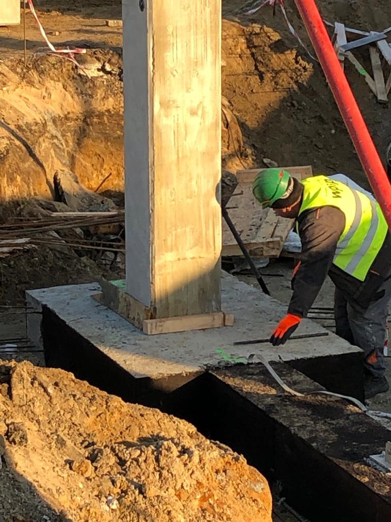 Pracownik w kasku i kamizelce odblaskowej przy betonowym słupie na fundamencie, przygotowujący zbrojenie, widok na wykop i teren budowy w słoneczny dzień.