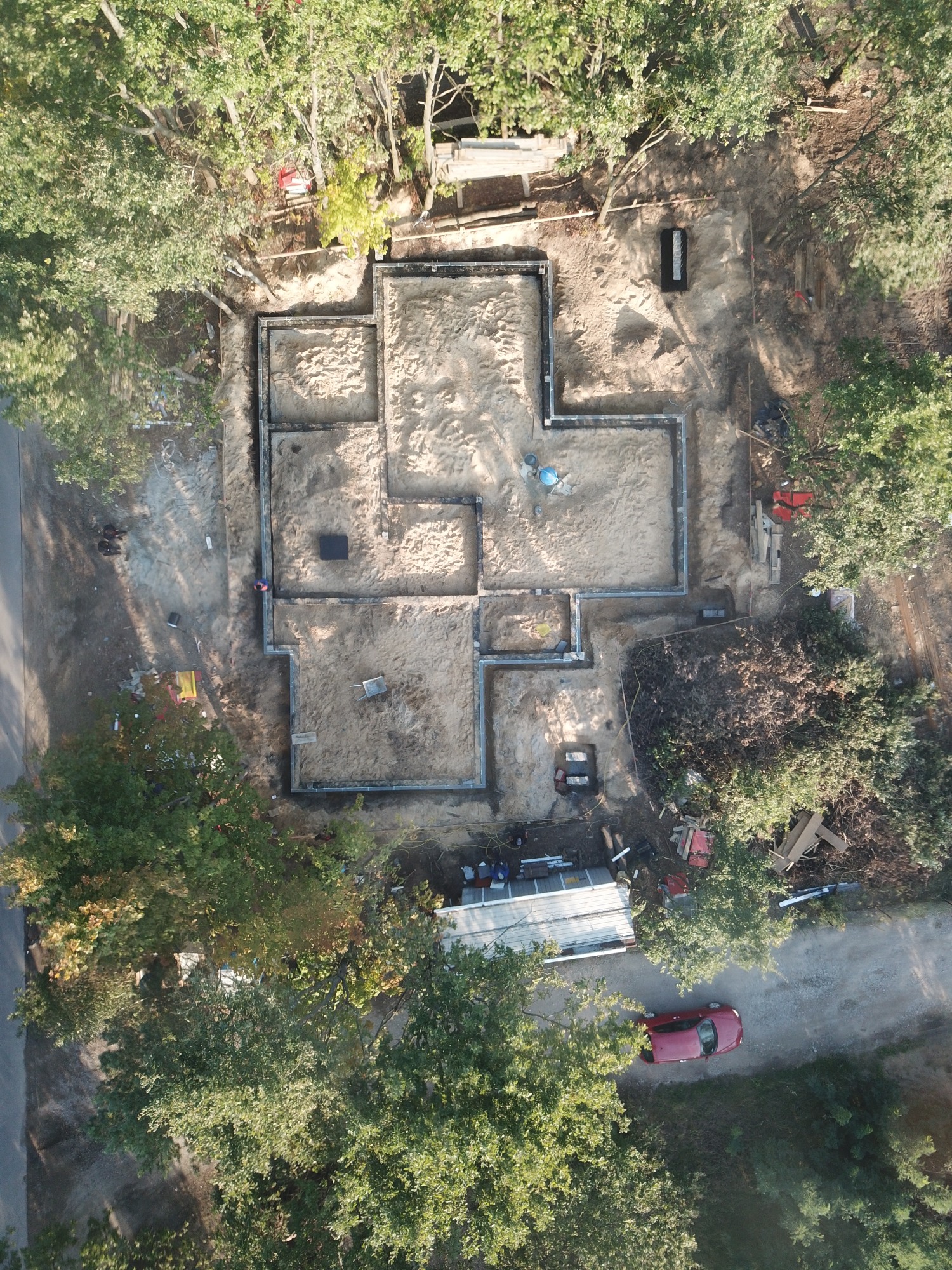 Widok z drona na plac budowy z wykopanymi fundamentami pod budynek, otoczony drzewami, z widocznym czerwonym samochodem na drodze dojazdowej.