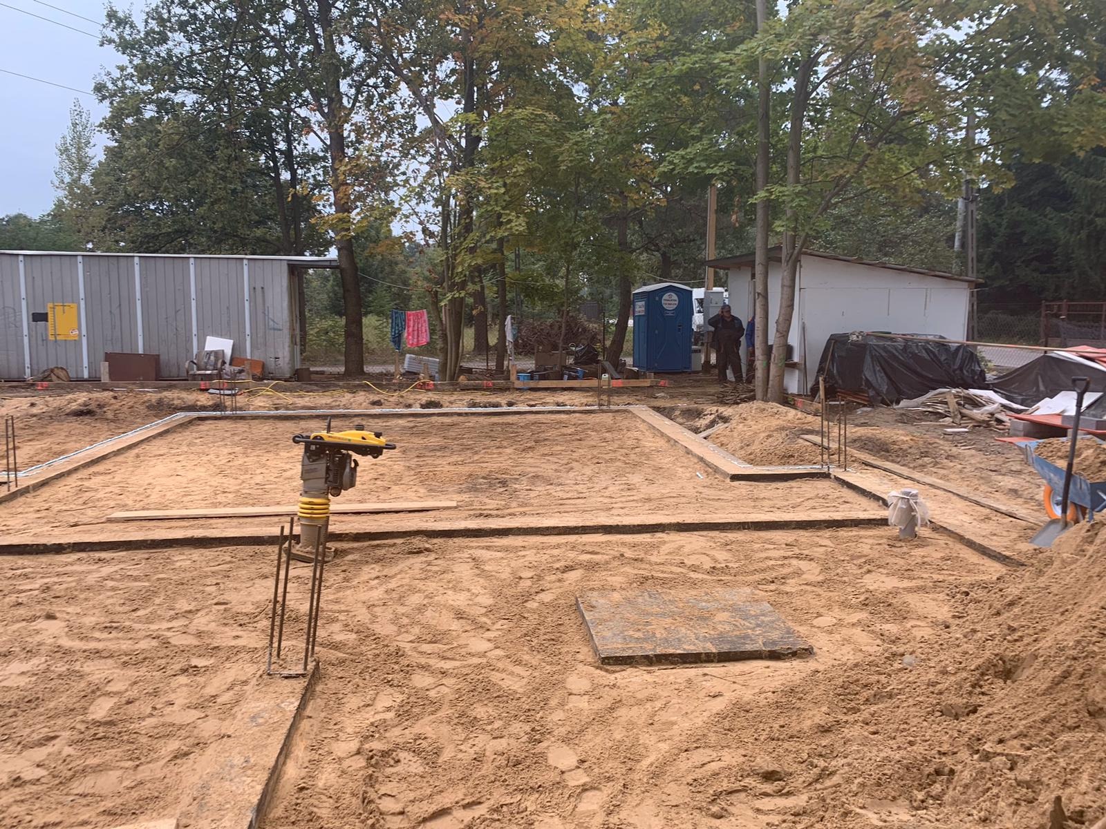 Przygotowanie terenu pod budowę domu: wykopane fundamenty, ubijarka wibrująca i wystające pręty zbrojeniowe, w tle budynek gospodarczy i przenośna toaleta.