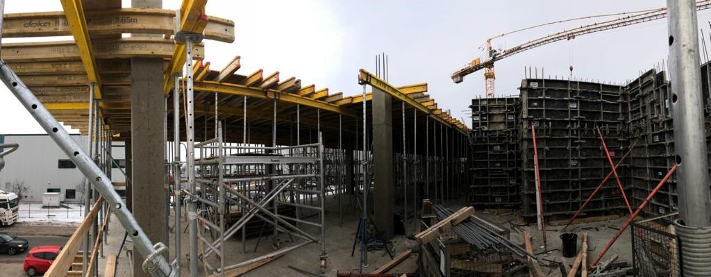 Szeroki kadr na plac budowy z widocznymi szalunkami stropowymi Doka, stalowymi rusztowaniami, elementami zbrojenia i żurawiem w tle, uchwycony w pochmurny dzień.