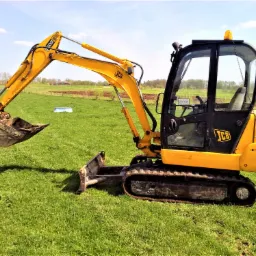 Żółto-czarna mini koparka JCB na zielonej trawie, z widocznym łyżką i lemieszem, w tle pola uprawne i zabudowania.