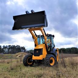 BIT-BUD Łukasz Bitowski - Żółto-czarna koparko-ładowarka JCB z podniesioną łyżką na tle zachmurzonego nieba i zalesionego krajobrazu.