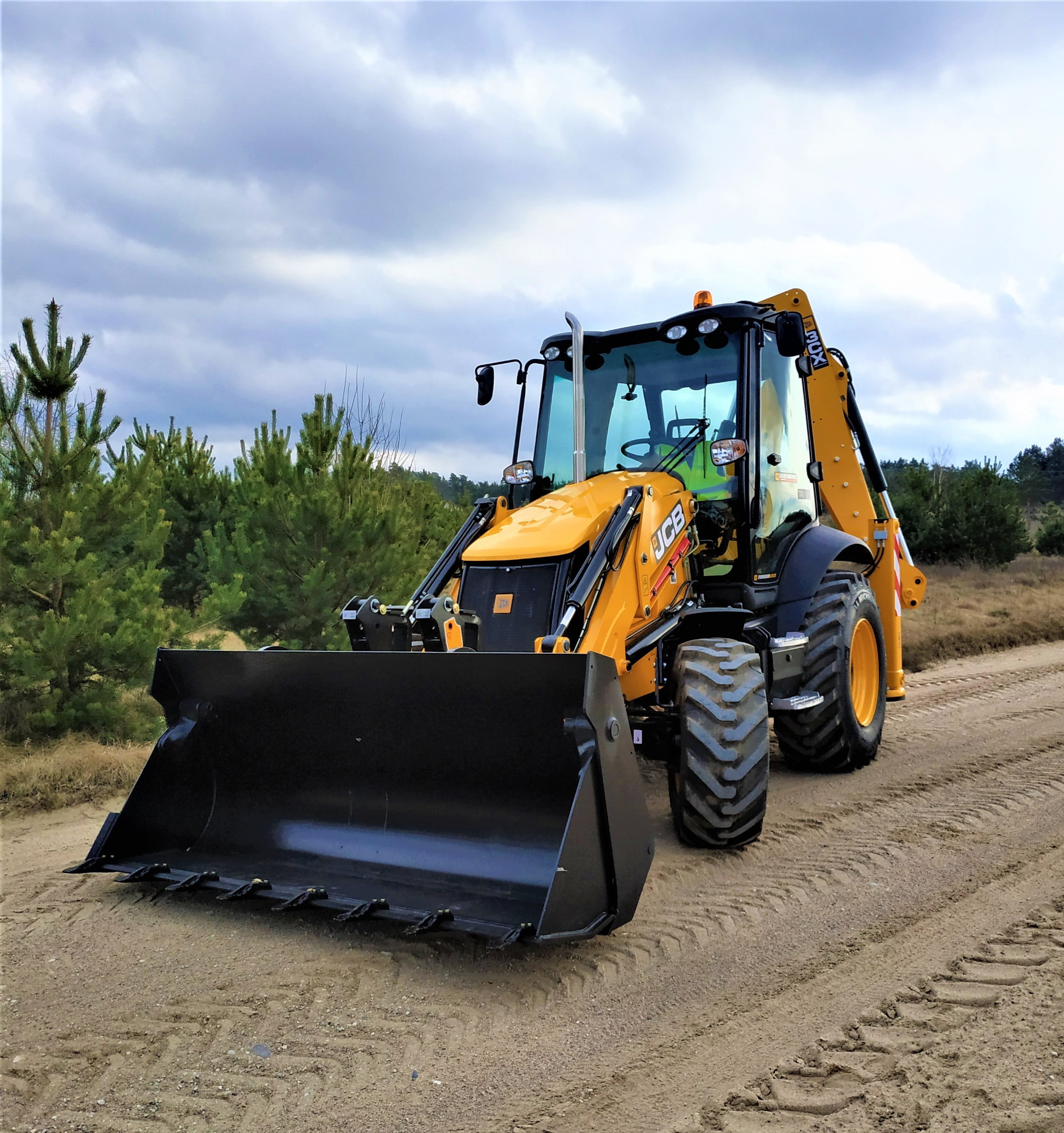 Żółto-czarna koparko-ładowarka JCB z opuszczoną łyżką na piaszczystej drodze, w tle las iglasty i pochmurne niebo.