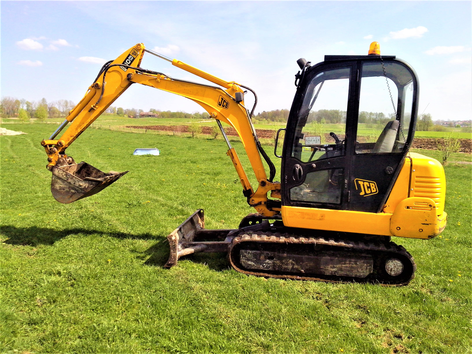Żółto-czarna mini koparka JCB na zielonej trawie, z widocznym łyżką i lemieszem, w tle pola uprawne i zabudowania.