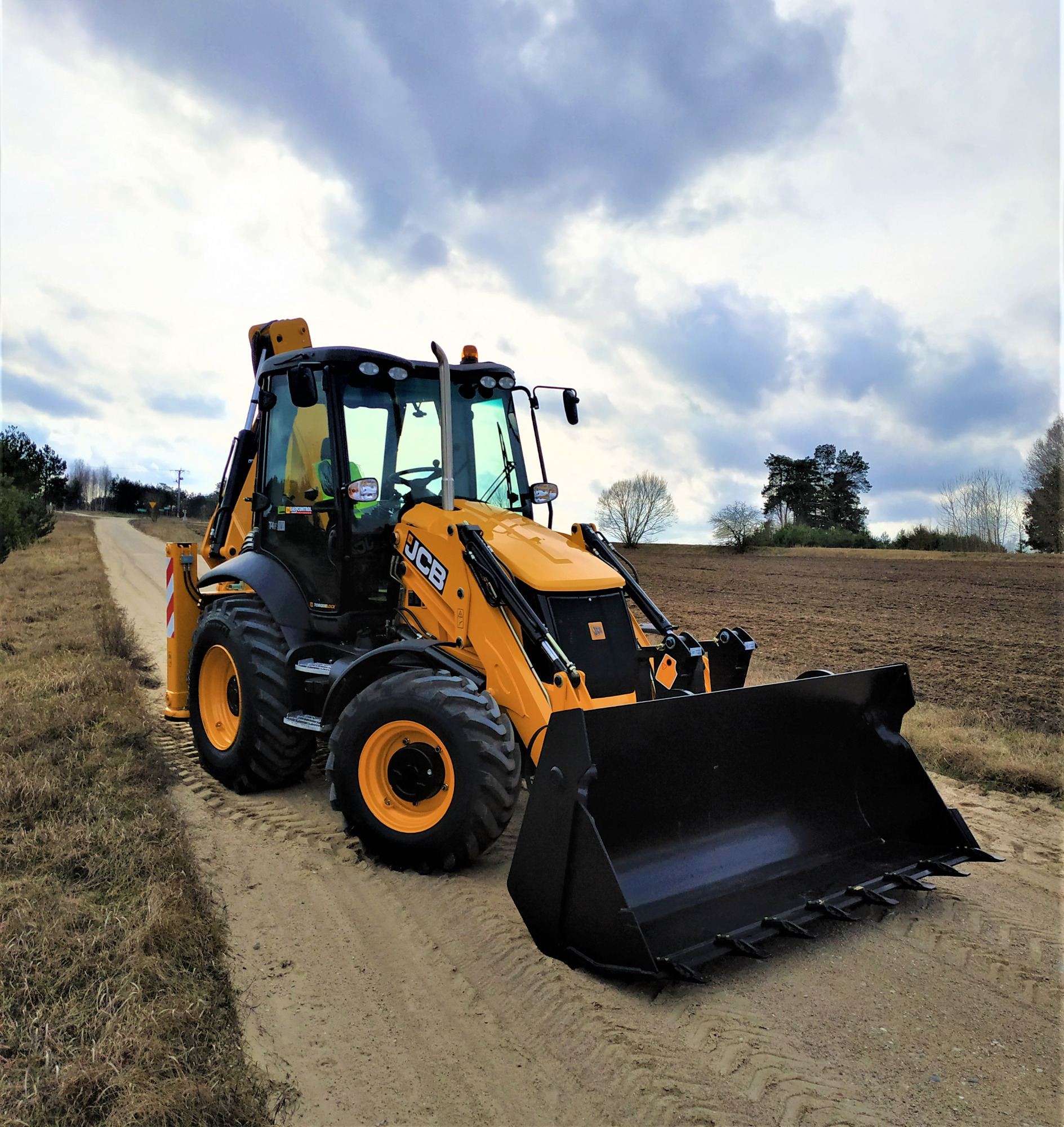 Żółto-czarna koparko-ładowarka JCB zaparkowana na piaszczystej drodze polnej, z widocznym polem i pochmurnym niebem w tle.