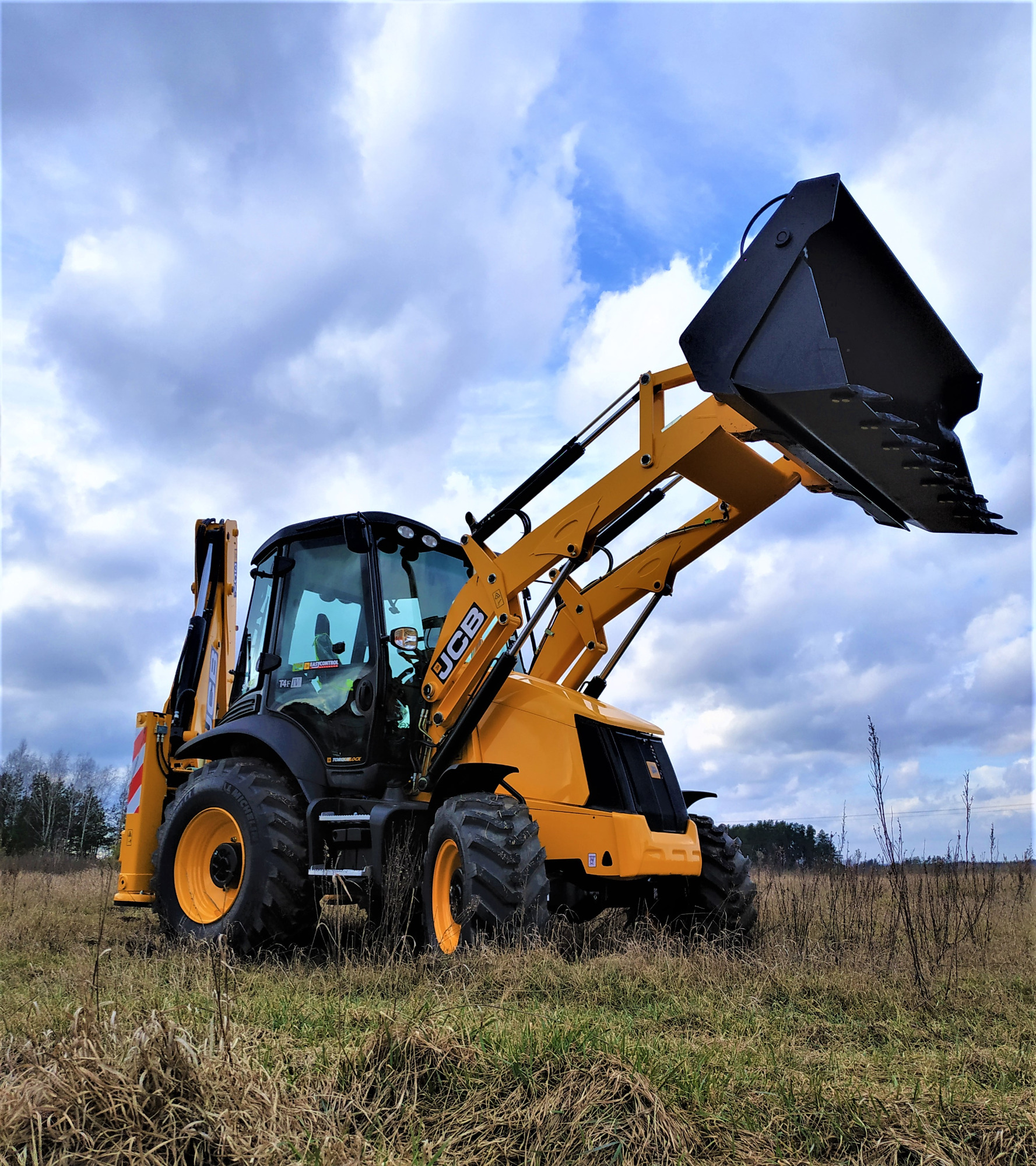 Żółto-czarna koparko-ładowarka JCB na polu ze skoszonymi trawami, z podniesioną łyżką, na tle pochmurnego nieba.
