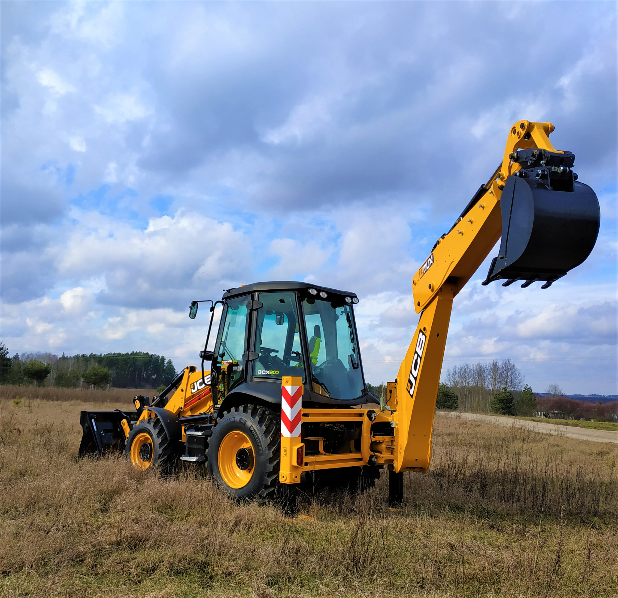 Żółto-czarna koparko-ładowarka JCB 3CXeco na polu z suchą trawą, z podniesionym ramieniem i łyżką, na tle pochmurnego nieba.