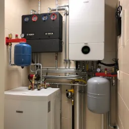 Zestaw urządzeń grzewczych marki Bosch w kotłowni, obejmujący kaskadę dwóch kotłów, zasobnik, naczynia przeponowe oraz liczne rury i zawory, wszystko zamontowane na tle beżowych płytek.